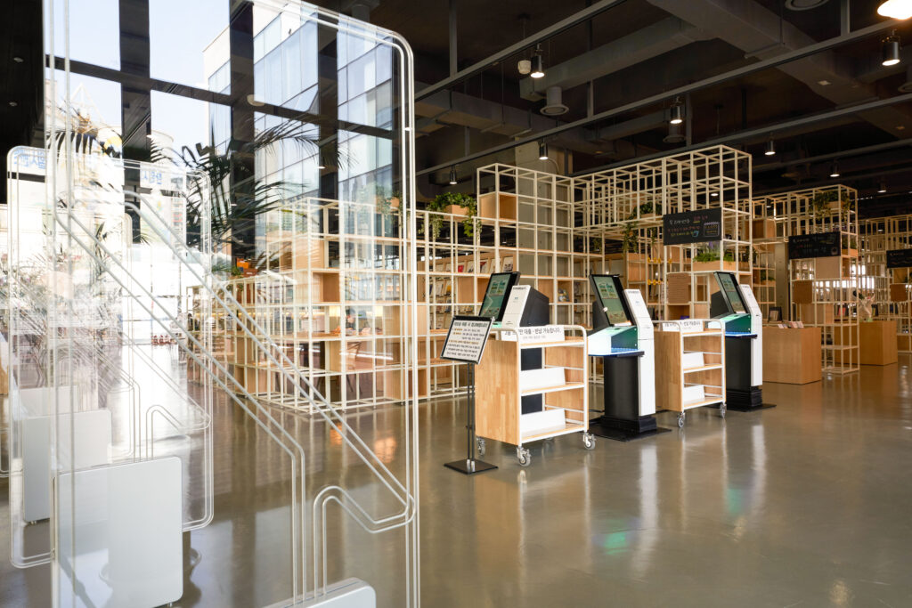 Interior de uma biblioteca moderna com espaço amplo, iluminação natural, estantes vazadas e totens de autoatendimento para empréstimo de livros e portão de segurança.