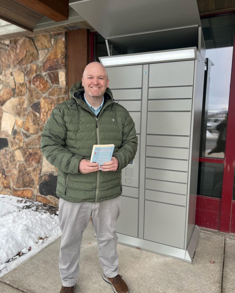 Summit County Library Director Dan Compton standing in front of the remoteLocker unit, smiling and holding a book on a snowy day
