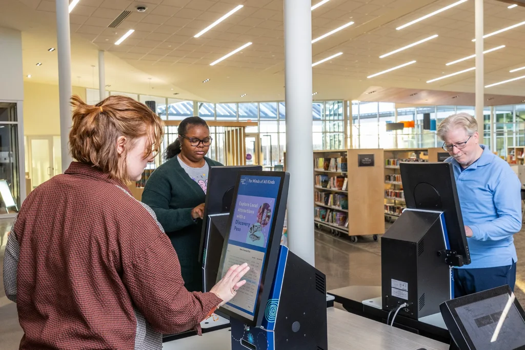 Three patrons using Bibliotheca selfCheck kiosks to view library events