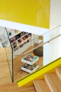 Vue intérieure moderne de la médiathèque B612 avec rayonnages de livres jeunesse et escalier aux finitions bois et jaune.